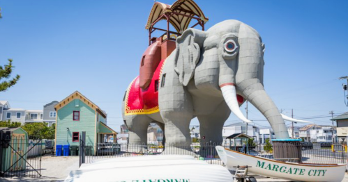 A large, whimsical elephant-shaped building with a red saddle, surrounded by a fence and nearby boats in Margate City.