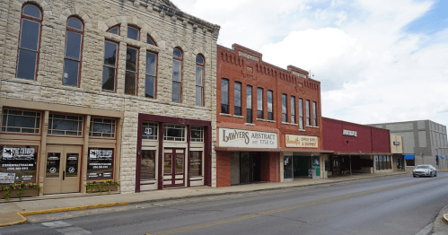 Historic buildings line a street, featuring stone and brick facades with storefronts and a clear blue sky above.