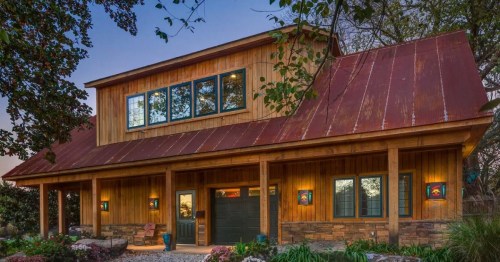 A rustic wooden house with a red metal roof, large windows, and a welcoming porch surrounded by greenery and flowers.