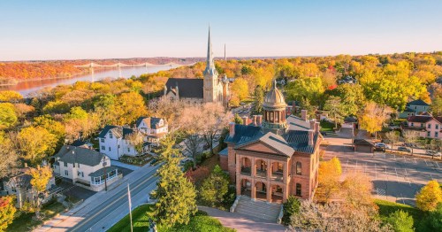 Aerial view of a town with historic buildings, a church, and colorful autumn trees by a river.