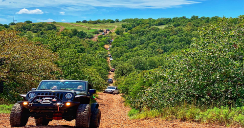 A line of off-road vehicles driving down a dirt path through a lush, green landscape under a blue sky.