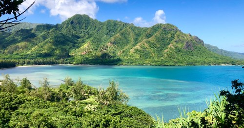 Lush green mountains rise above a tranquil blue bay, with clear skies and gentle waves lapping at the shore.