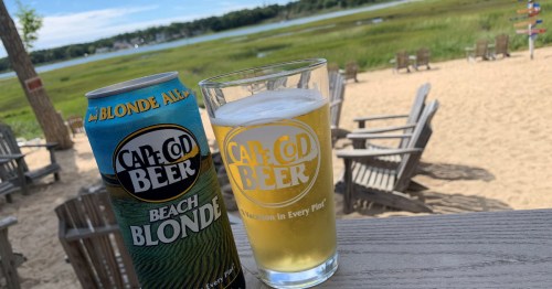 A can and glass of Cape Cod Beach Blonde beer on a wooden table by a sandy beach with chairs and a scenic view.