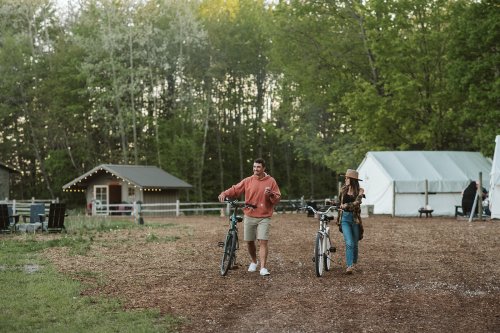 A man and woman walk with bicycles in a grassy area, surrounded by trees and tents, enjoying a sunny day outdoors.