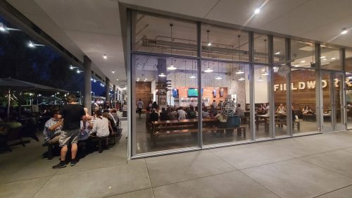 A lively restaurant scene at night, with diners inside and outside, illuminated by warm lights and a modern design.