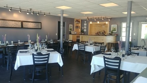A modern restaurant interior with white tablecloths, black chairs, and a bar in the background. Soft lighting enhances the ambiance.
