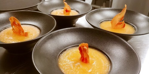 Four black bowls of orange soup, each topped with a crispy, curled piece of prosciutto.