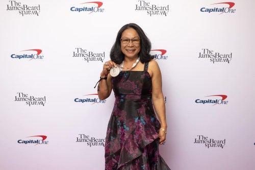 A woman in a floral dress holds a medal, smiling at a red carpet event with James Beard and Capital One logos in the background.