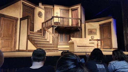 A theater stage set with a wooden staircase, a couch, and several doors, ready for a performance. Audience in foreground.