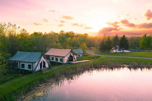A serene sunset over glamping tents by a calm pond, surrounded by lush greenery and trees.