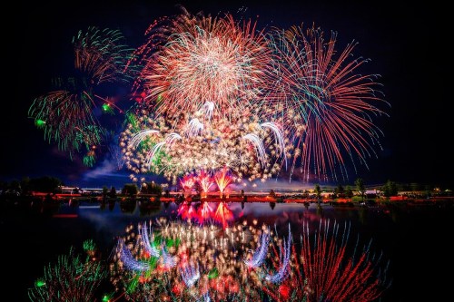 Colorful fireworks explode in the night sky, reflecting on a calm lake below.