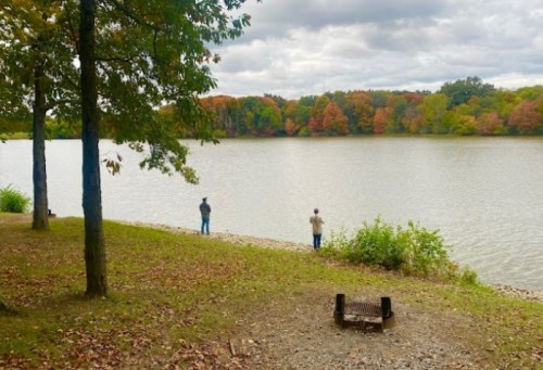 Two people fish by a calm lake surrounded by trees with autumn foliage under a cloudy sky.