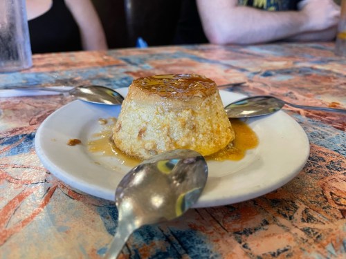 A creamy flan dessert on a white plate, surrounded by four spoons, on a colorful table.