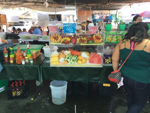 A vibrant fruit stand at a market, featuring colorful fruits and snacks, with people dining in the background.