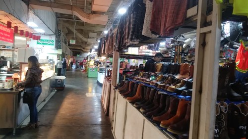 A bustling market scene with stalls selling boots and food, shoppers browsing in a vibrant indoor space.