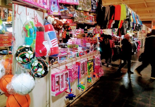 A vibrant market stall filled with colorful toys, sports equipment, and clothing, with shoppers browsing nearby.