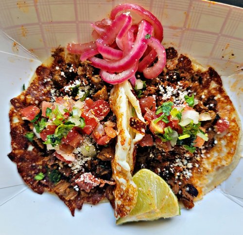 Two tacos filled with meat, topped with diced tomatoes, onions, cilantro, cheese, pickled onions, and a lime wedge.