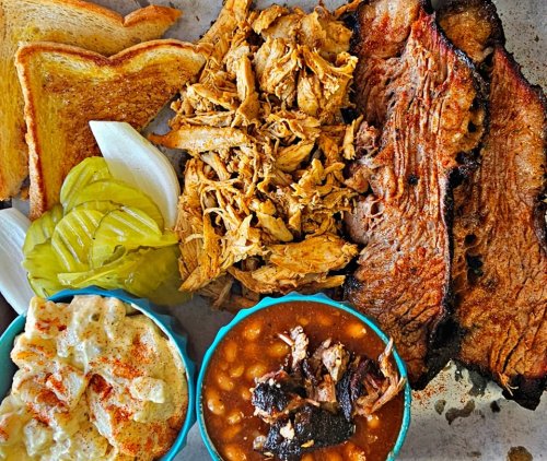 A platter of barbecue featuring sliced brisket, pulled chicken, pickles, onions, toast, and sides of baked beans and potato salad.