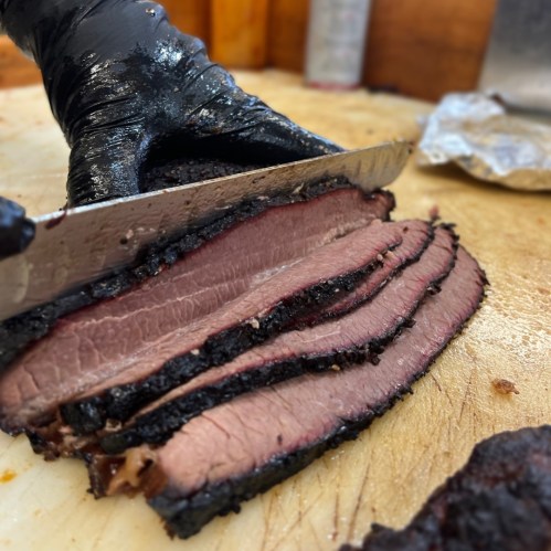 A close-up of a hand in a black glove slicing smoked brisket on a wooden cutting board.