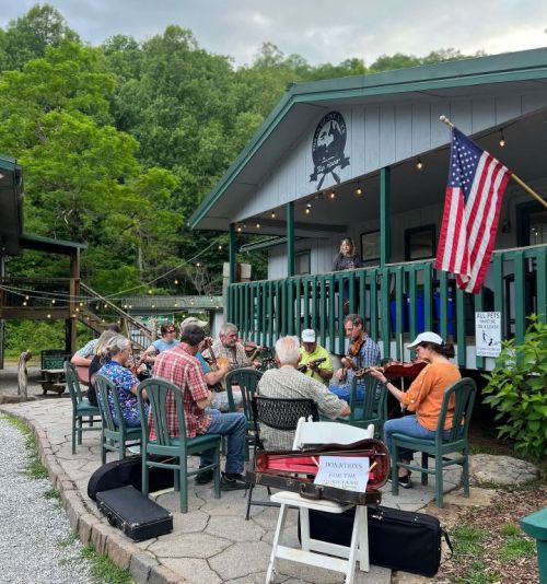 A group of musicians playing instruments outdoors on a patio, with an American flag and greenery in the background.