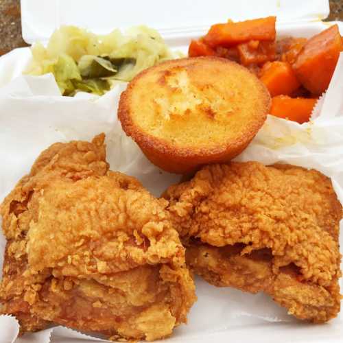 A plate of fried chicken, cornbread, sweet potatoes, and pickled vegetables in a takeout container.