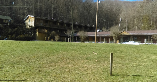 A rustic building with multiple levels, surrounded by trees and a grassy area, set against a mountainous backdrop.