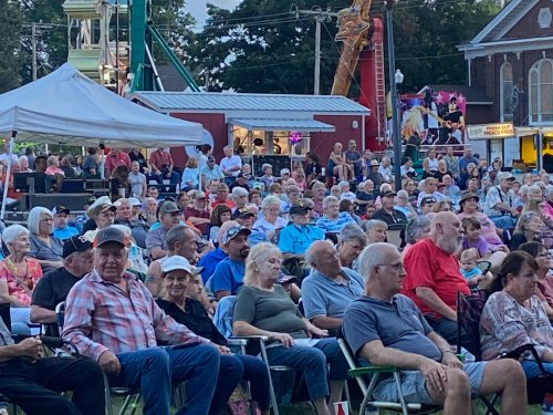 A large crowd of people seated outdoors at an event, with tents and amusement rides in the background.
