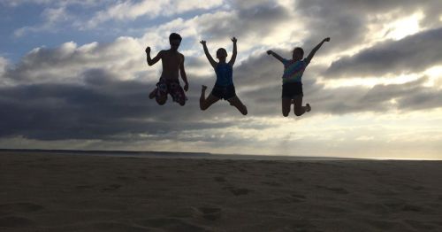 Three people jumping joyfully on a beach against a cloudy sky, with the sun setting in the background.