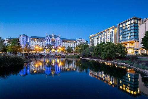 A hotel complex by a calm lake at dusk, with colorful lights reflecting on the water and trees surrounding the area.