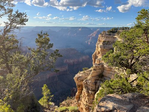 A scenic view of the Grand Canyon, featuring rocky cliffs, lush greenery, and a clear blue sky with scattered clouds.