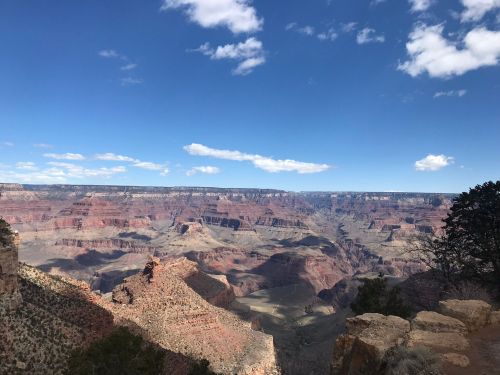 A panoramic view of the Grand Canyon, showcasing layered rock formations under a blue sky with scattered clouds.