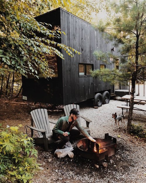 A person sits by a fire pit outside a black tiny house, surrounded by trees and a small dog nearby.