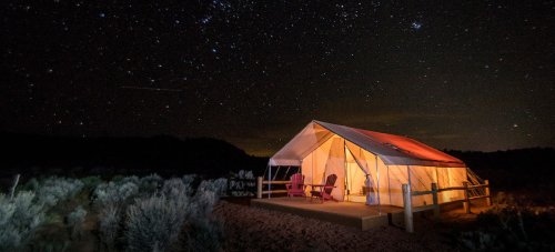 A glamping tent illuminated at night, surrounded by a starry sky and desert landscape.
