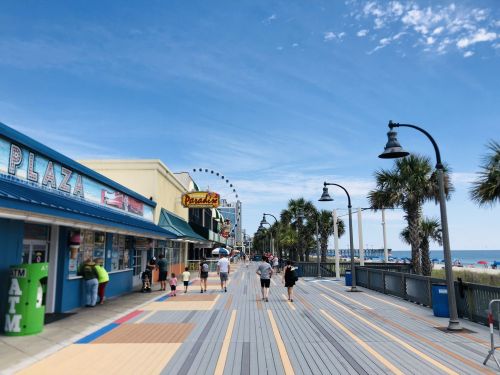 A sunny boardwalk lined with palm trees, shops, and a ferris wheel in the distance, with people strolling along.