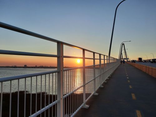 A sunset view from a bridge, with a pathway beside water and city skyline in the distance.
