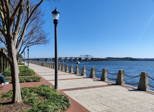 A scenic waterfront path lined with trees and lampposts, overlooking a river and bridge under a clear blue sky.