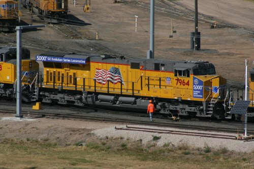 A yellow locomotive with an American flag design, parked on tracks with a worker in an orange vest nearby.