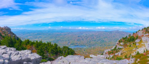 A panoramic view of a colorful valley surrounded by mountains and trees under a bright blue sky with scattered clouds.