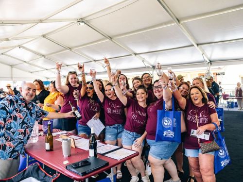 A group of smiling people in matching shirts raise glasses in celebration under a tent at an event.