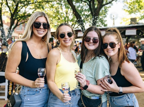 Four women in sunglasses smile and pose together, each holding a glass of wine, in a lively outdoor setting.