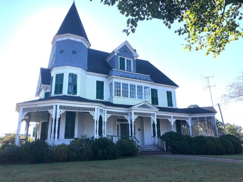 A large, historic white house with a pointed tower, green shutters, and a wraparound porch, set in a grassy area.