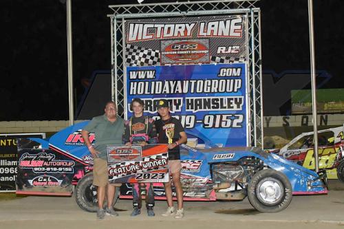 A driver stands with two supporters beside a race car, celebrating a feature win in Victory Lane.