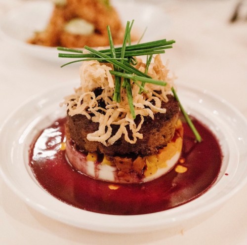 A plated dish featuring a layered cake topped with crispy onions and chives, surrounded by a rich sauce.