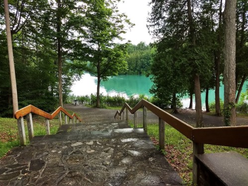 A stone staircase leads down to a tranquil, turquoise lake surrounded by lush green trees.