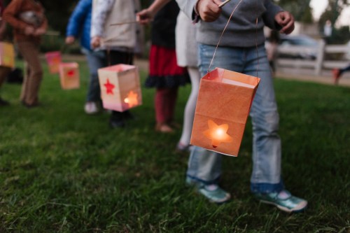 Children walking on grass, holding glowing paper lanterns with star cutouts during an evening event.