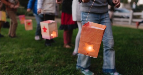A group of people holding colorful paper lanterns with glowing lights, walking on grass during an evening event.