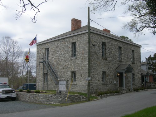 Historic stone building with a Maryland flag, surrounded by trees and a gravel road, under a cloudy sky.