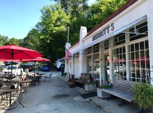 A quaint storefront with outdoor seating, red umbrellas, and American flags, surrounded by greenery.