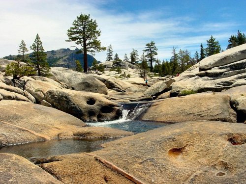 A serene landscape featuring smooth rocks, a small waterfall, and pine trees under a clear blue sky.