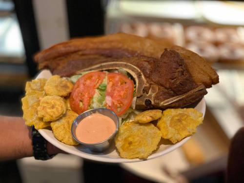 A plate of crispy pork belly, sliced tomatoes, lettuce, and fried plantains served with a dipping sauce.